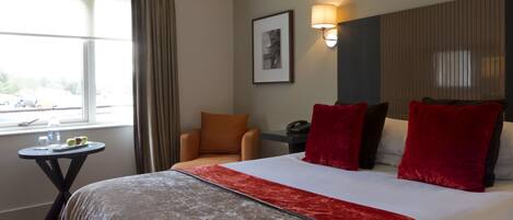 Habitación de hotel con una cama grande, una mesita con botella de agua y fruta, una silla y una ventana con cortinas.