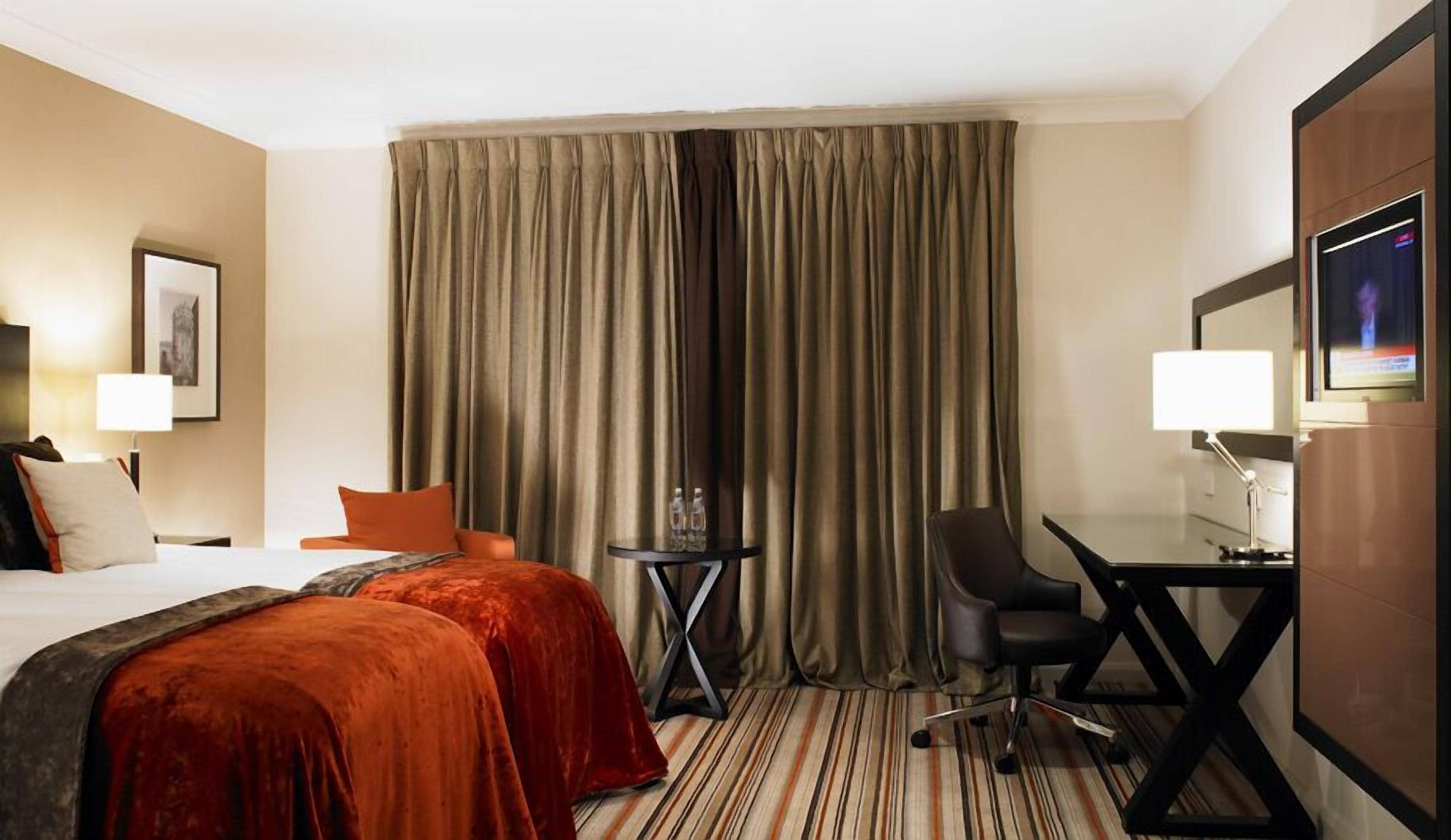 A hotel room with a bed, two bedside tables, a desk with a chair, a TV, and a large window with curtains.