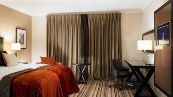 Une chambre d’hôtel comprenant un lit, deux tables de chevet, un bureau avec une chaise, une télévision et une grande fenêtre avec des rideaux.