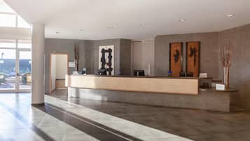 Uma área de recepção de hotel moderna, com um balcão de atendimento extenso, amplas janelas e piso em azulejos.