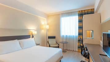 Habitación de hotel con una cama grande, un escritorio con computadora, una silla, una mesita y una lámpara.