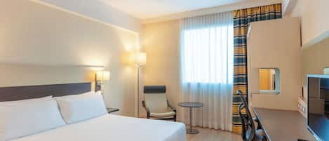 A hotel room with a large bed, a desk with a computer, a chair, a small table, and a lamp.