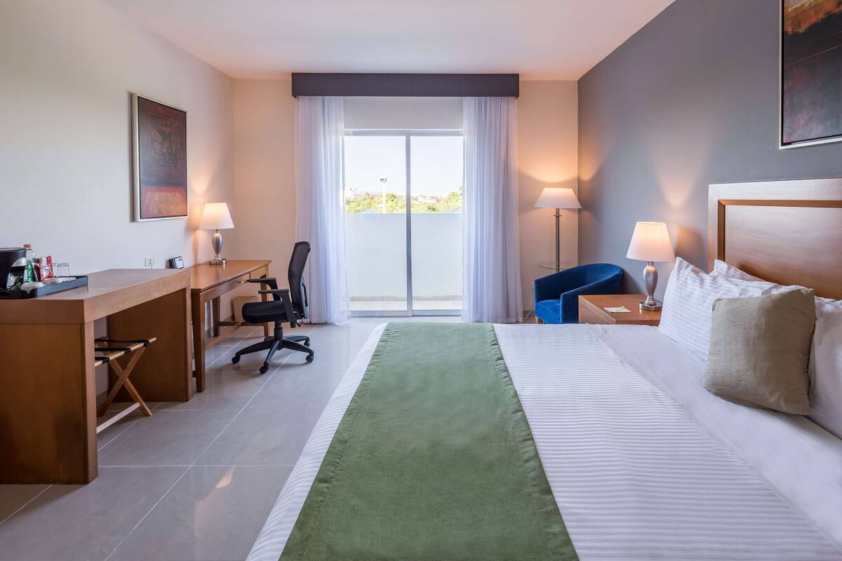 A hotel room with a bed, desk, chair, and a view of the outdoors through a large window.