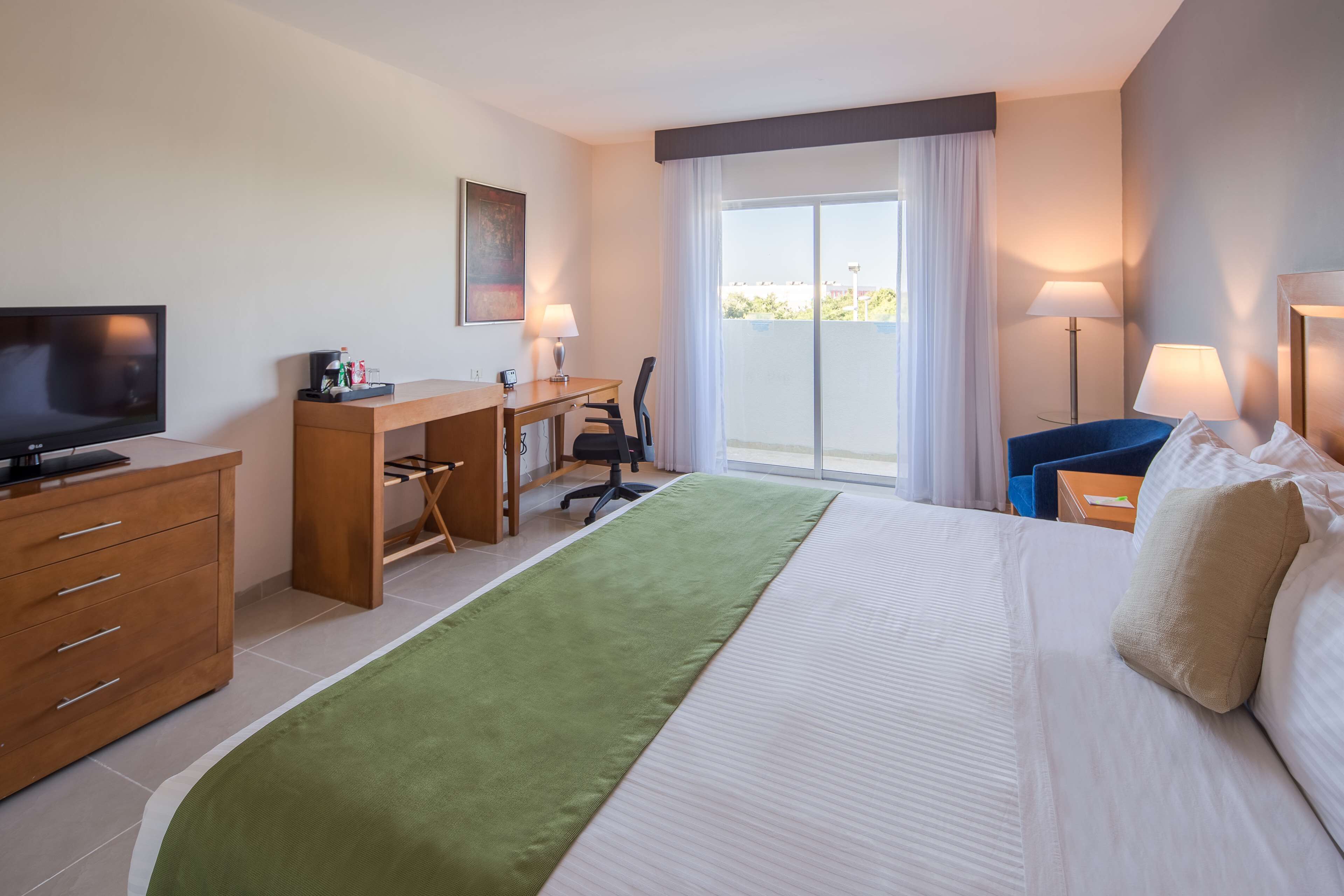 Habitación de hotel con cama, escritorio, silla, televisor y balcón con vistas.
