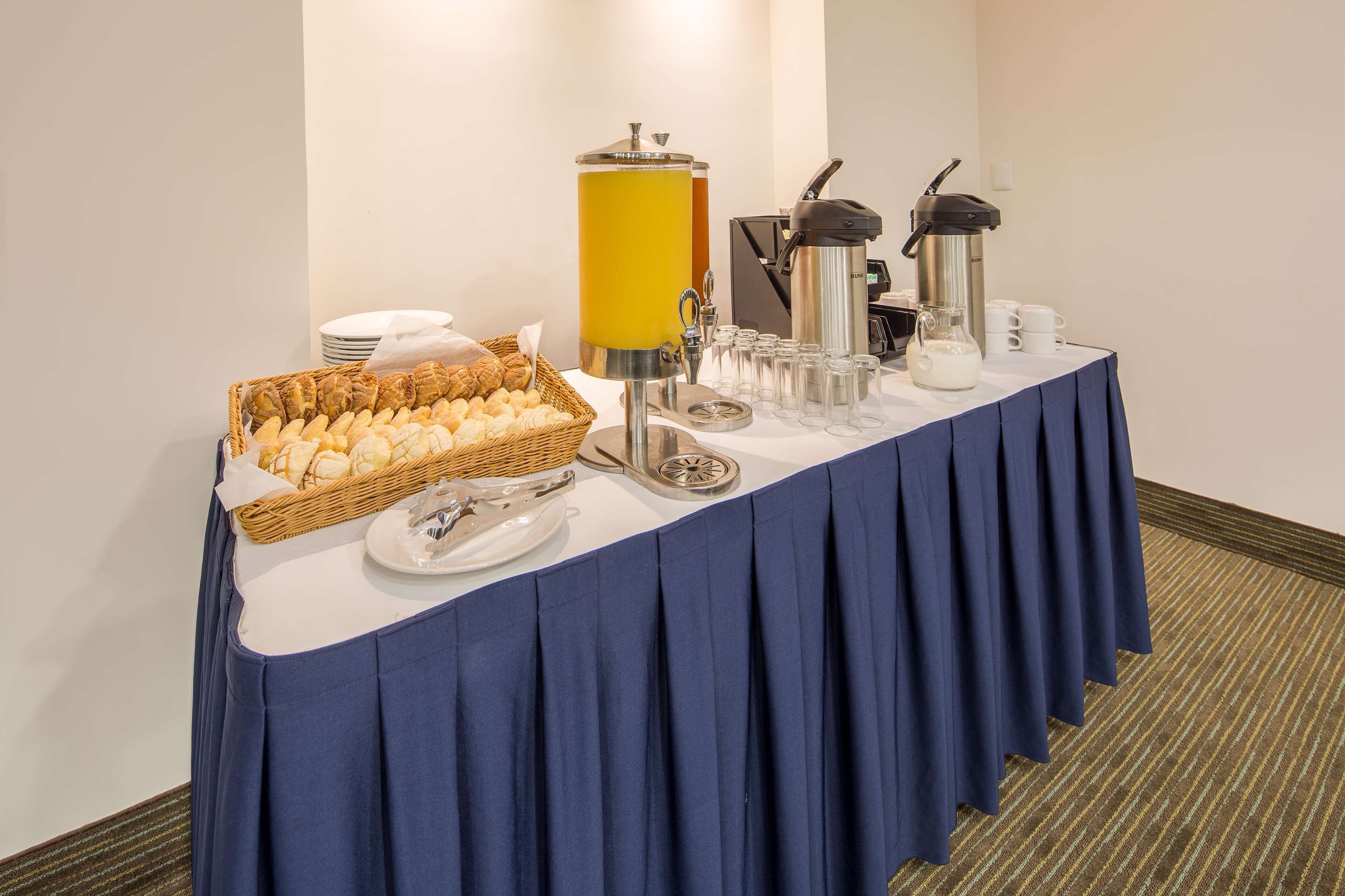 Una mesa de buffet con mantel azul, una cesta de pan, una máquina de café y un dispensador de zumo.