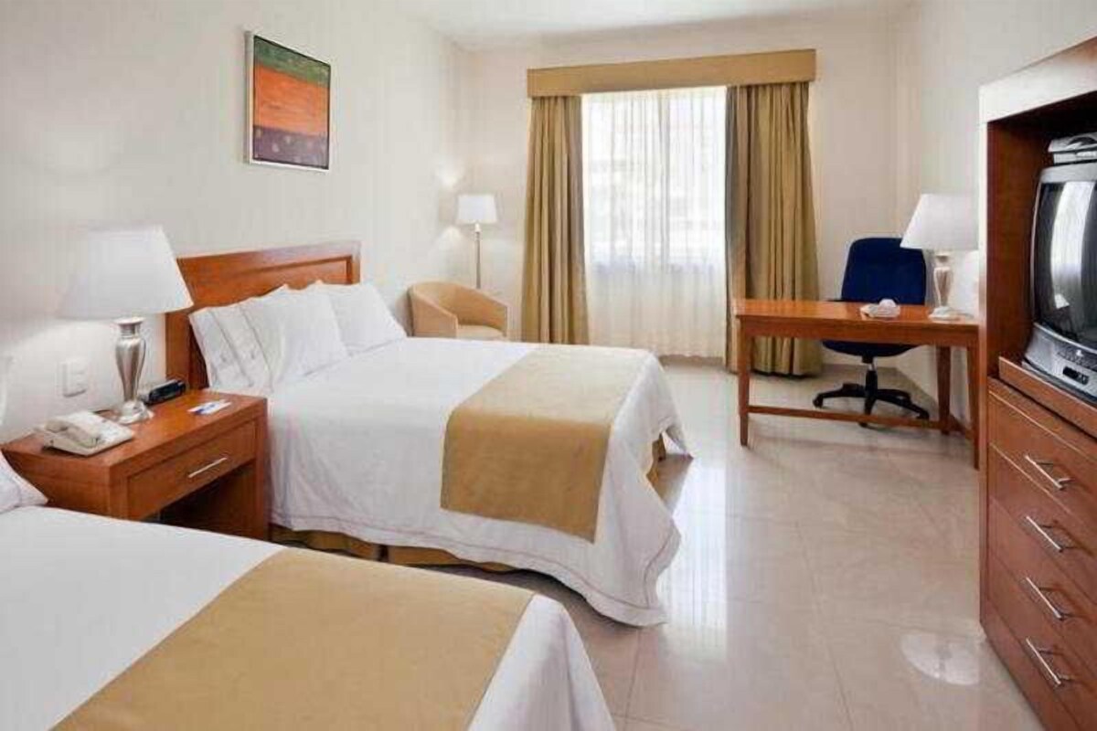 A hotel room with two beds, a desk, a chair, a television, and a window with curtains.