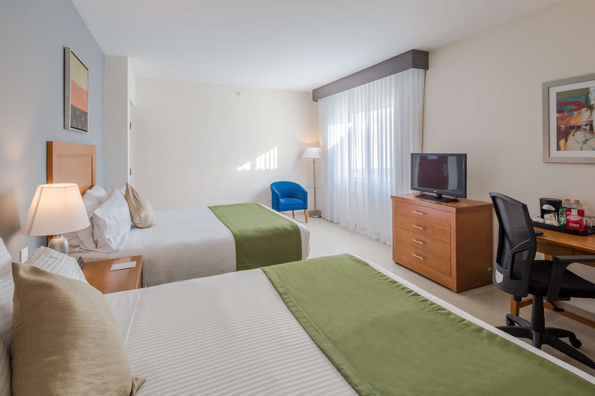 A hotel room with two beds, a desk with a computer, a chair, a TV, and a lamp.