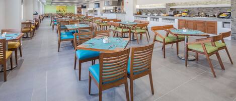 A spacious dining area with multiple tables and chairs, a kitchen area with a buffet, and a large flat-screen TV mounted on the wall.