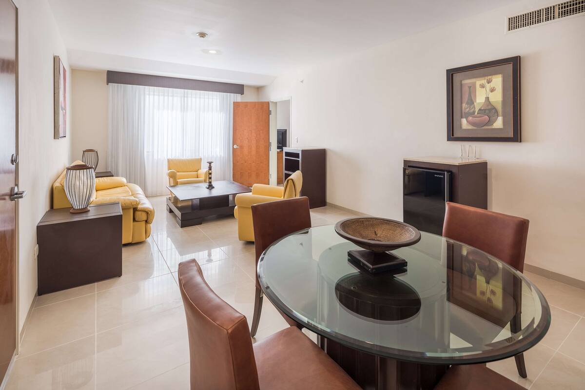 A modern living room with a glass-top dining table, leather sofas, and a large window with curtains.