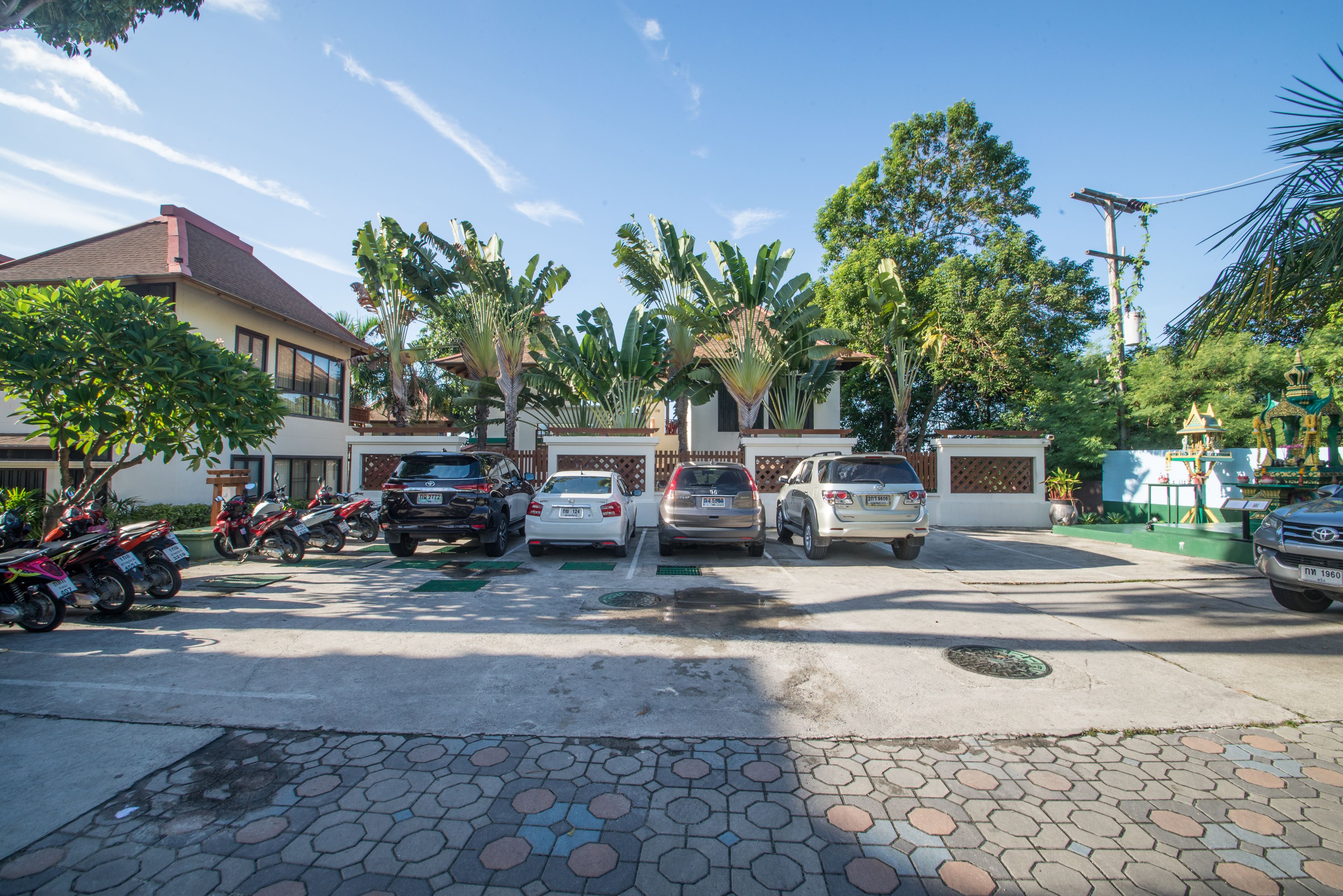 Um estacionamento com vários carros e motocicletas, rodeado por árvores e um edifício com telhado vermelho.