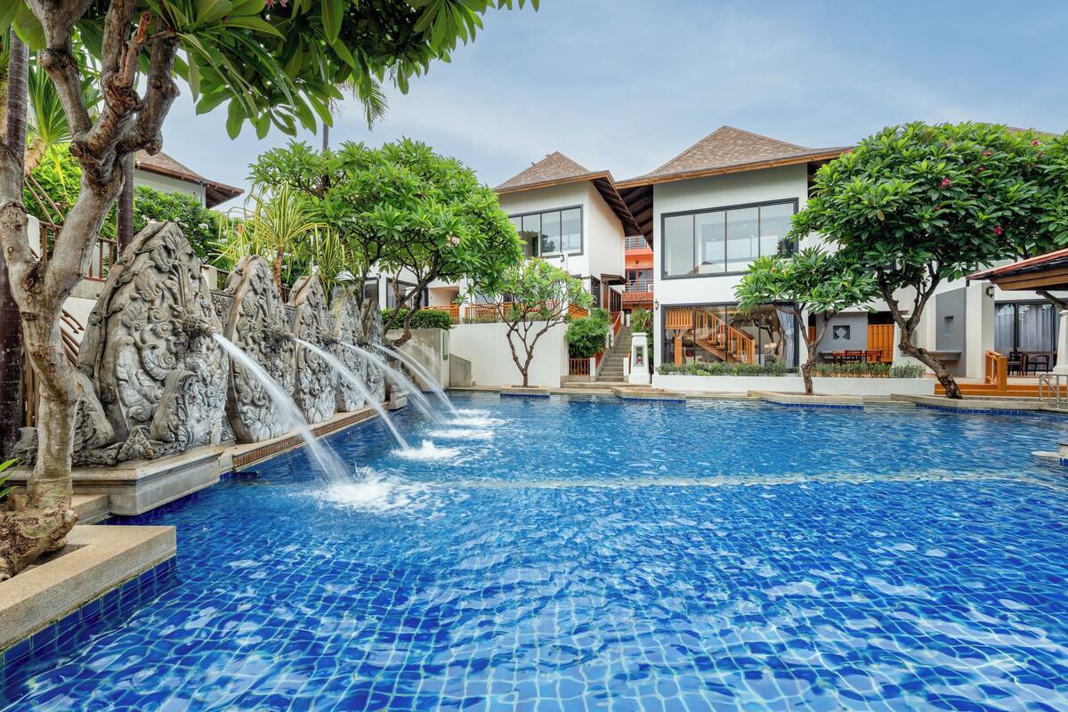 A swimming pool with a waterfall feature, surrounded by modern buildings and lush greenery.