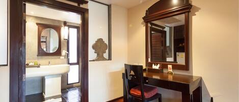 A hotel room with a wooden desk, a chair, a large mirror, and a view of the bathroom.