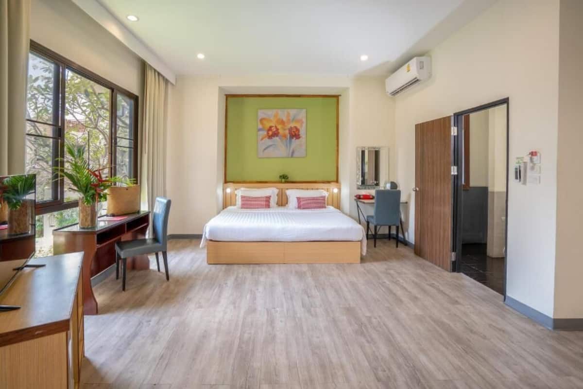 A modern hotel room with a large bed, a desk, and a chair. There is a window with plants, a painting on the wall, and an air conditioning unit.