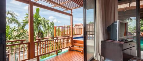 A balcony with wooden flooring, a TV on a cabinet, and a view of palm trees and a pool.