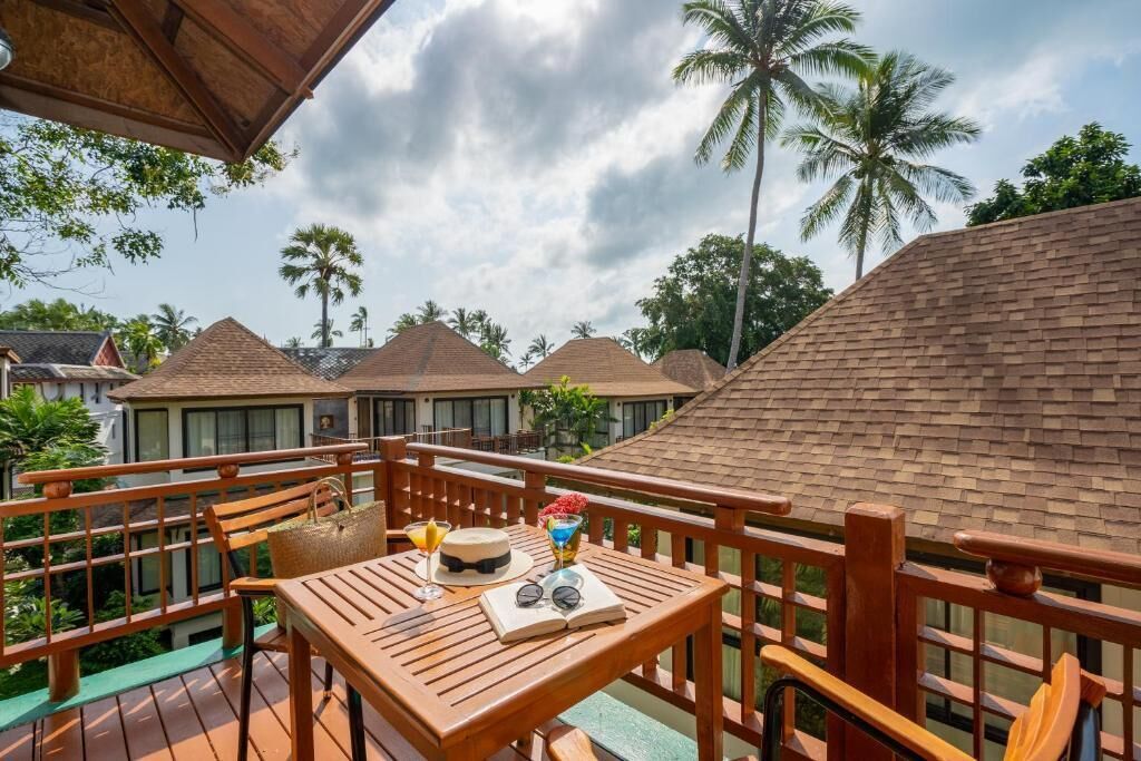 A wooden deck with a table set for two, overlooking a resort with palm trees and thatched-roof buildings.