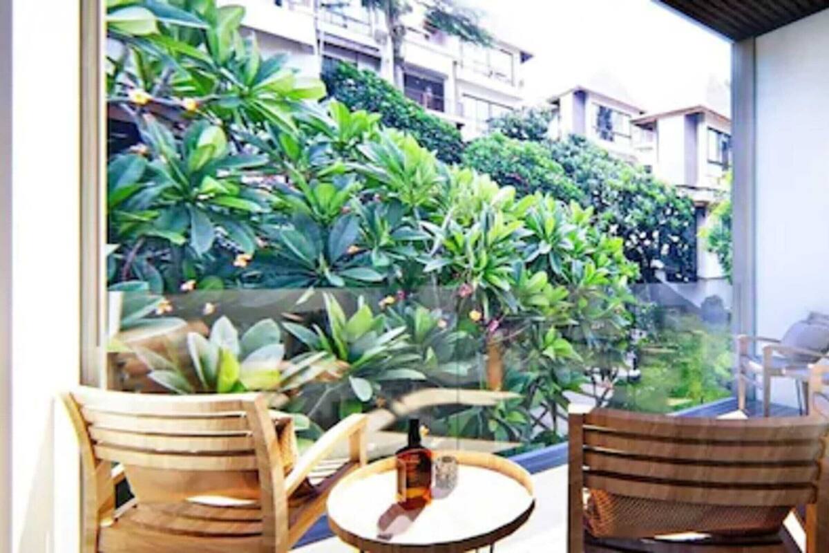 A balcony with wooden chairs and a table, overlooking a lush green garden and residential buildings.