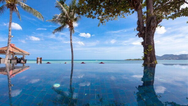 An infinity pool with a clear view of the ocean, surrounded by palm trees and a small building with a red roof. - The Briza Beach Resort, Samui (Koh Samui)