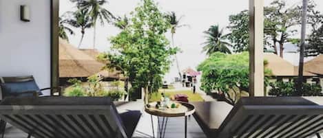 A modern outdoor seating area with two lounge chairs and a small round table, overlooking a tropical landscape with palm trees and a house in the distance.