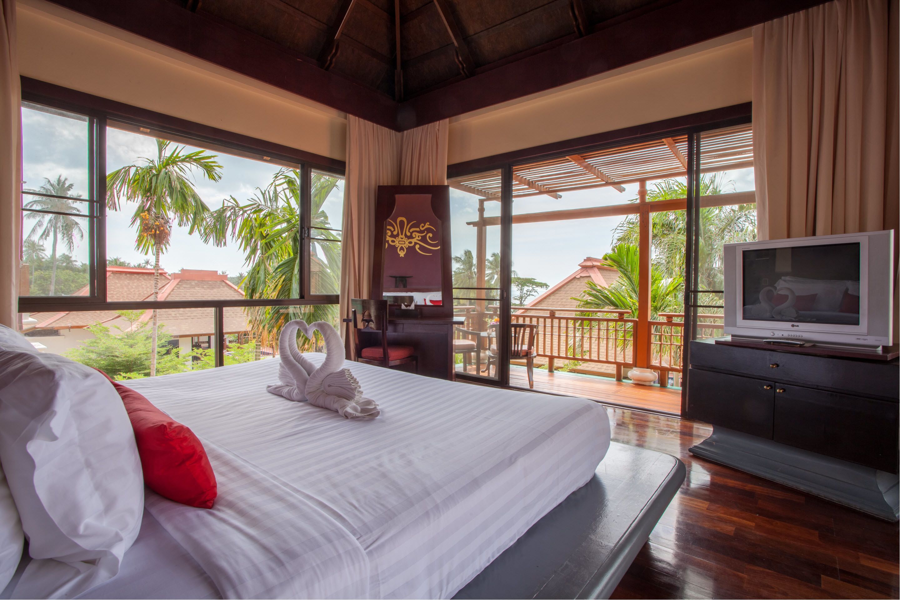 A hotel room with a large bed, a TV, and a balcony with a view of palm trees and a building.
