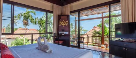 Habitación de hotel con una cama grande, un escritorio, un televisor y un balcón que ofrece vistas a palmeras y casas.