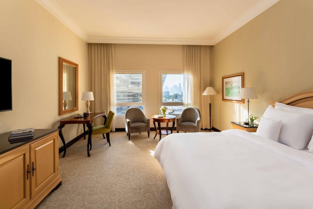 Ein Hotelzimmer mit einem Bett, zwei Sesseln, einem Schreibtisch, einem Fernseher und einem großen Fenster mit Blick auf die Stadt.