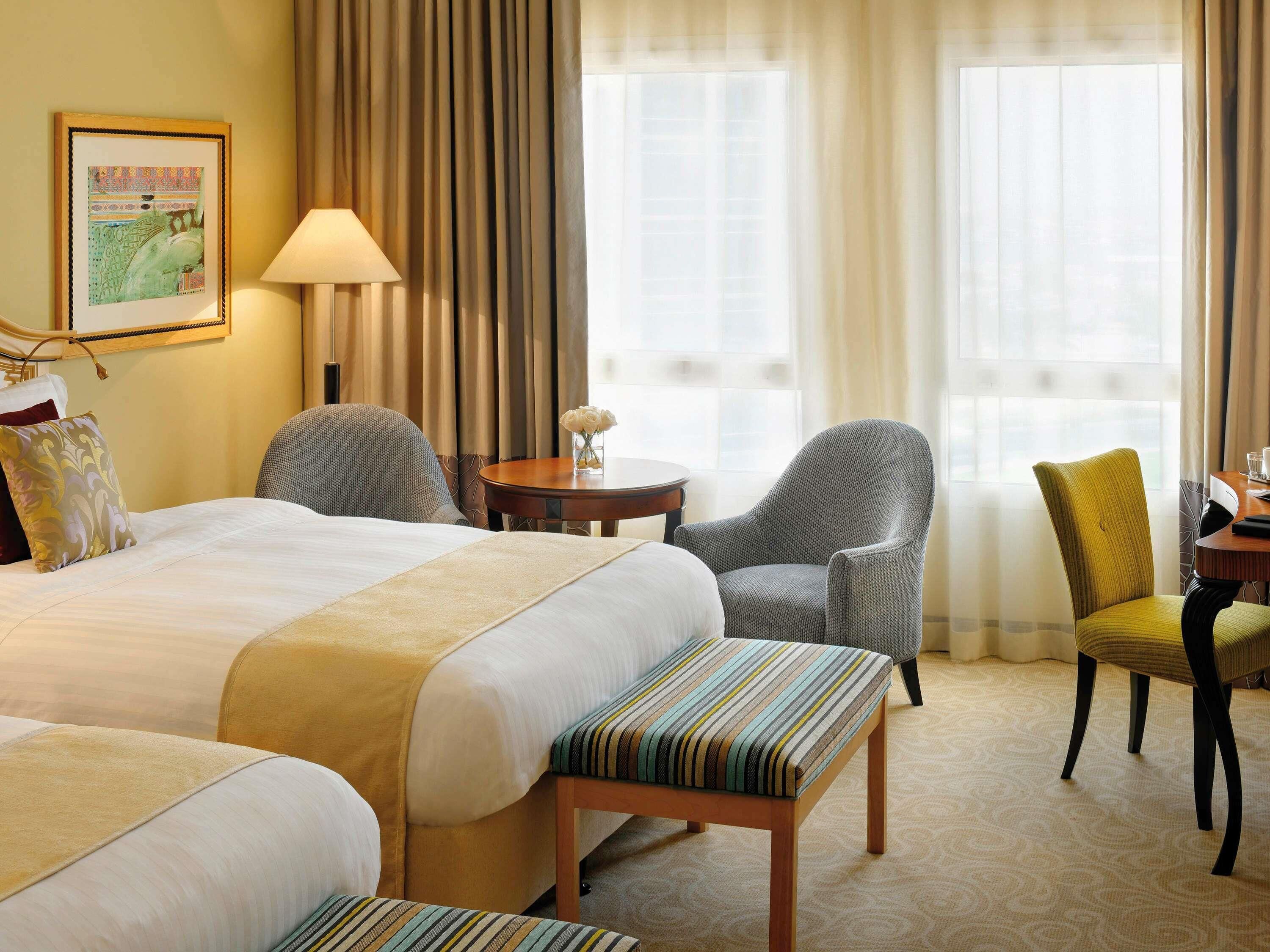 Una habitación de hotel con dos camas, una zona de estar con una silla y una mesa, y un ventanal con cortinas.