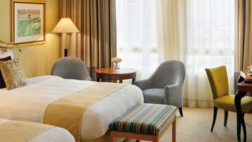 Una habitación de hotel con dos camas, una zona de estar con una silla y una mesa, y un ventanal con cortinas.