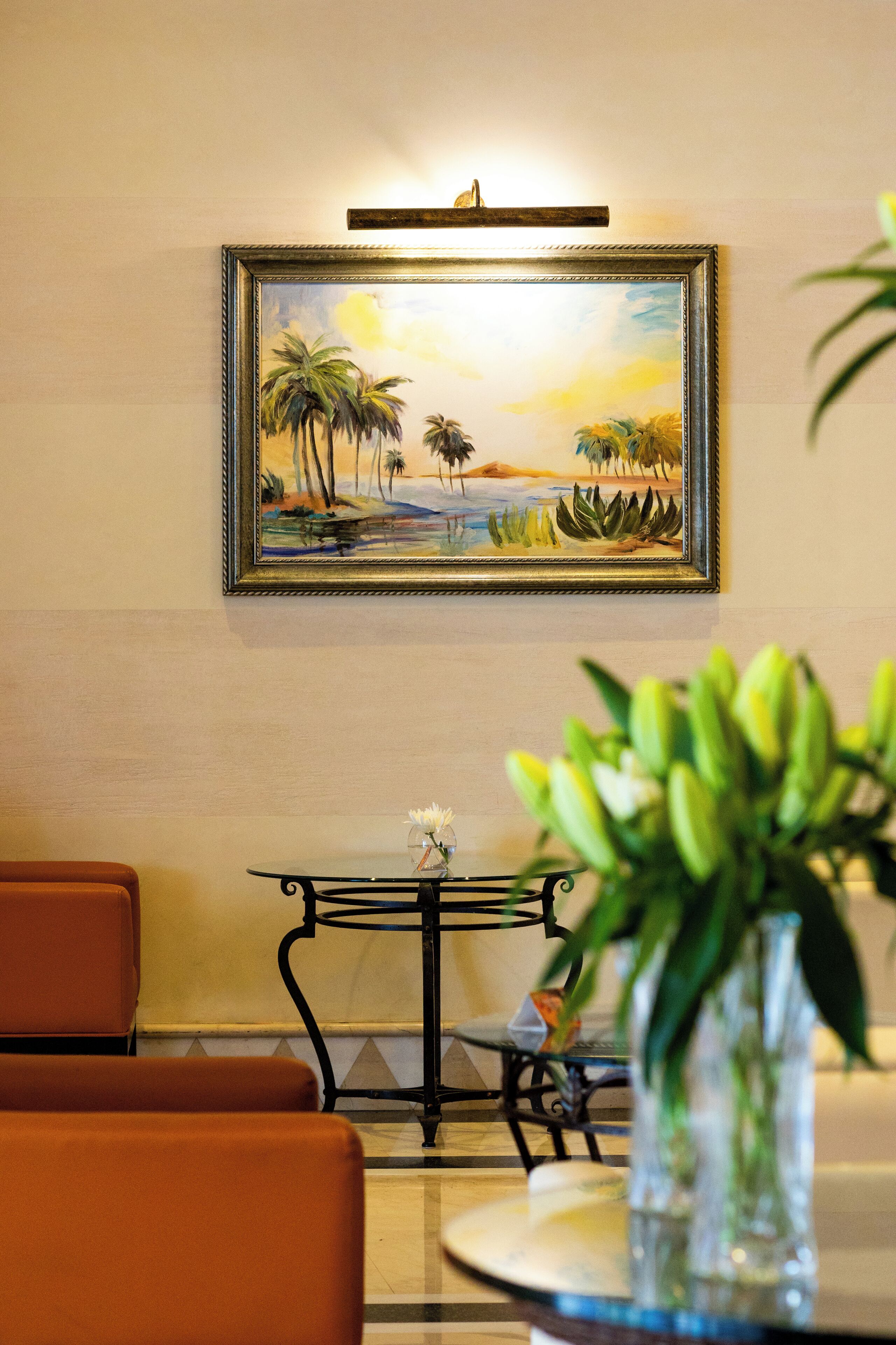 A framed painting of a tropical landscape on a wall, a small table with a vase of green plants, and orange chairs in a room.