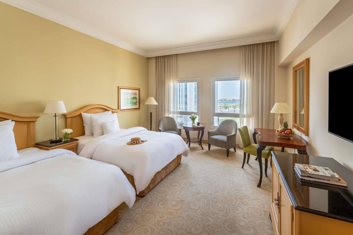 Ein Hotelzimmer mit zwei Betten, einem Schreibtisch mit Fernseher, einem Sessel und Blick auf die Stadt durch große Fenster.