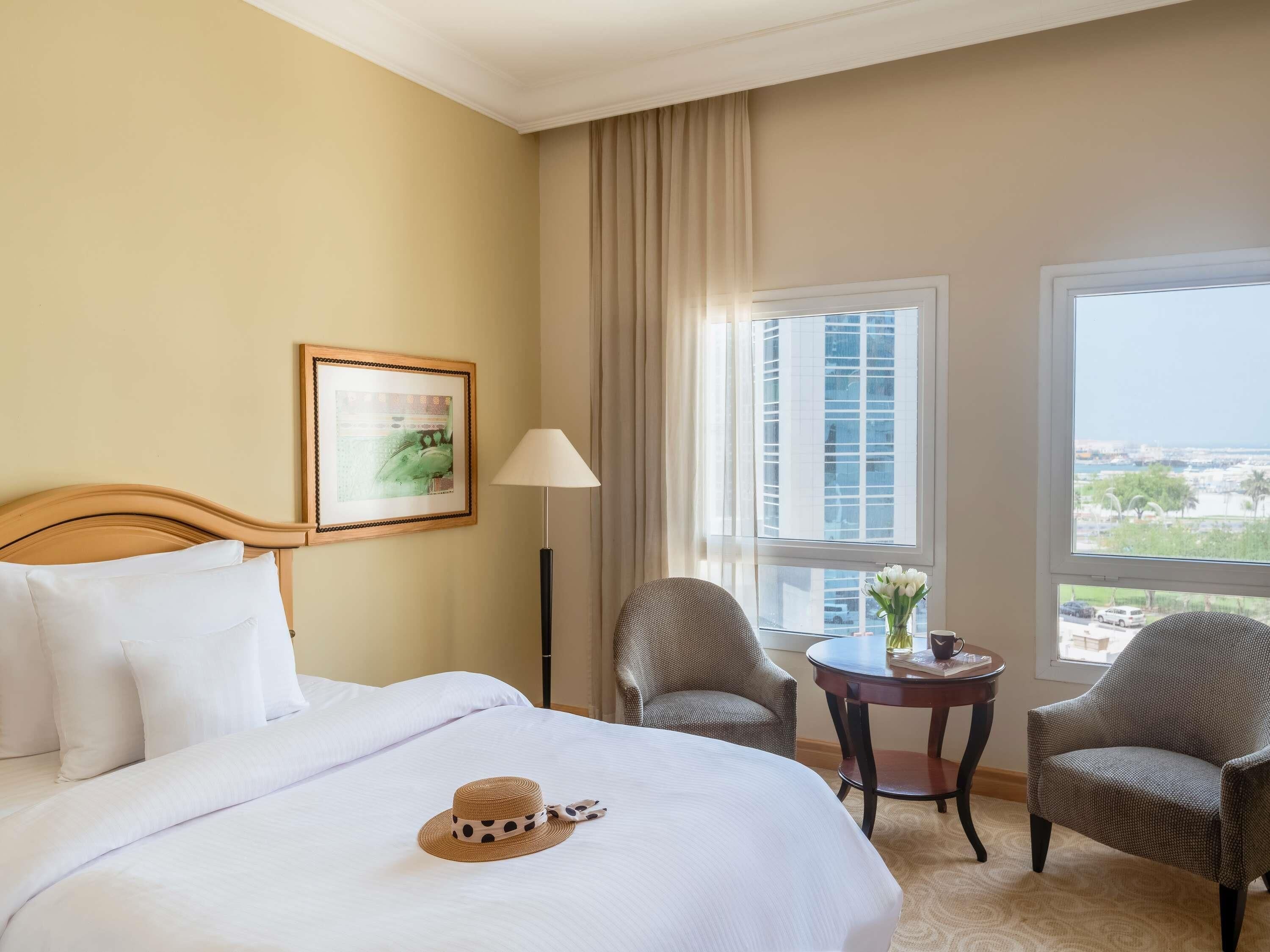 A hotel room with a bed, two armchairs, a small table with a vase, a lamp, and a window with a city view.