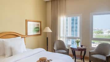 A hotel room with a bed, two armchairs, a small table with a vase, a lamp, and a window with a city view.