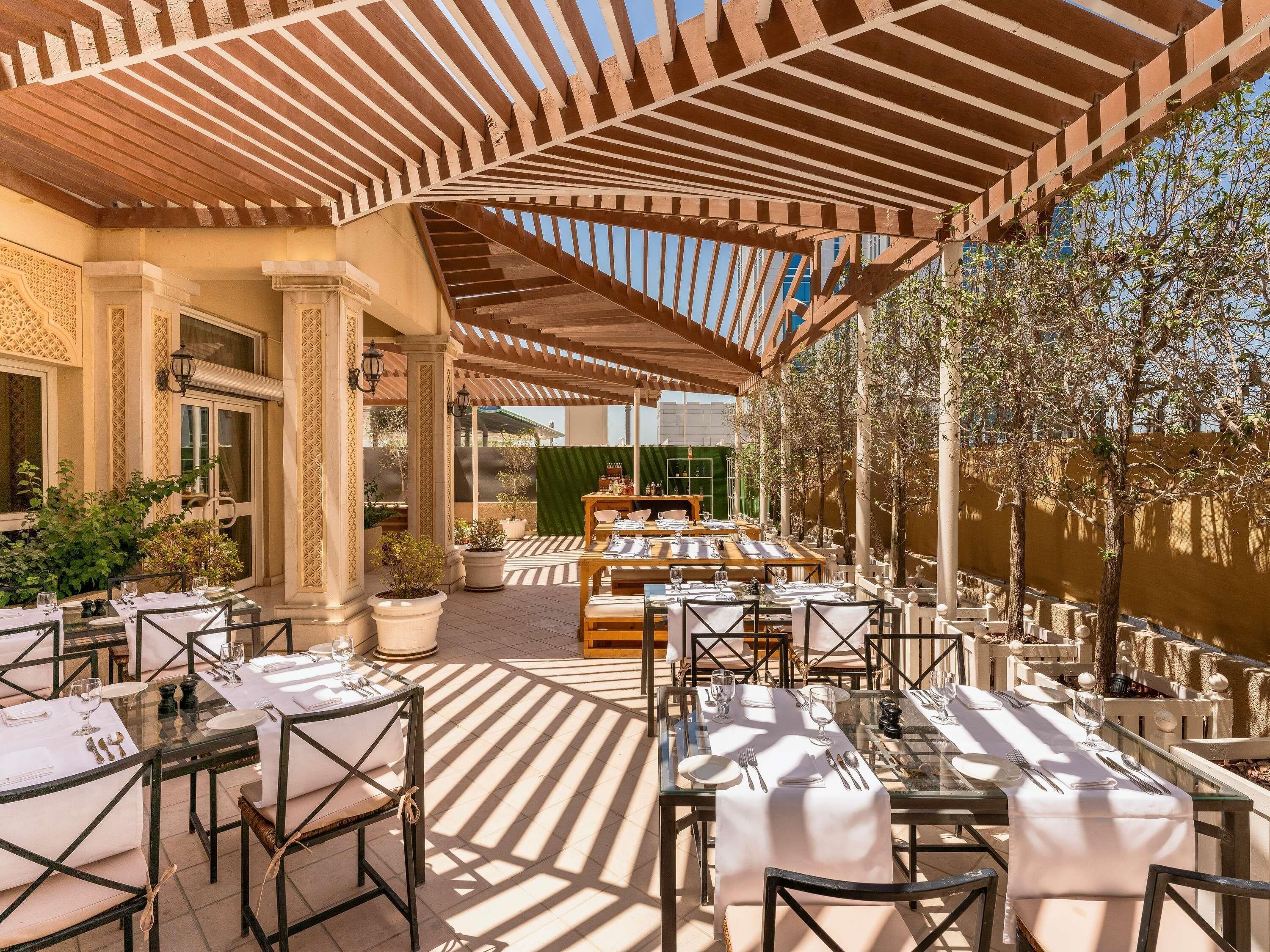 An outdoor dining area with tables set for two, covered by a wooden pergola, surrounded by potted plants and a view of a building in the background.