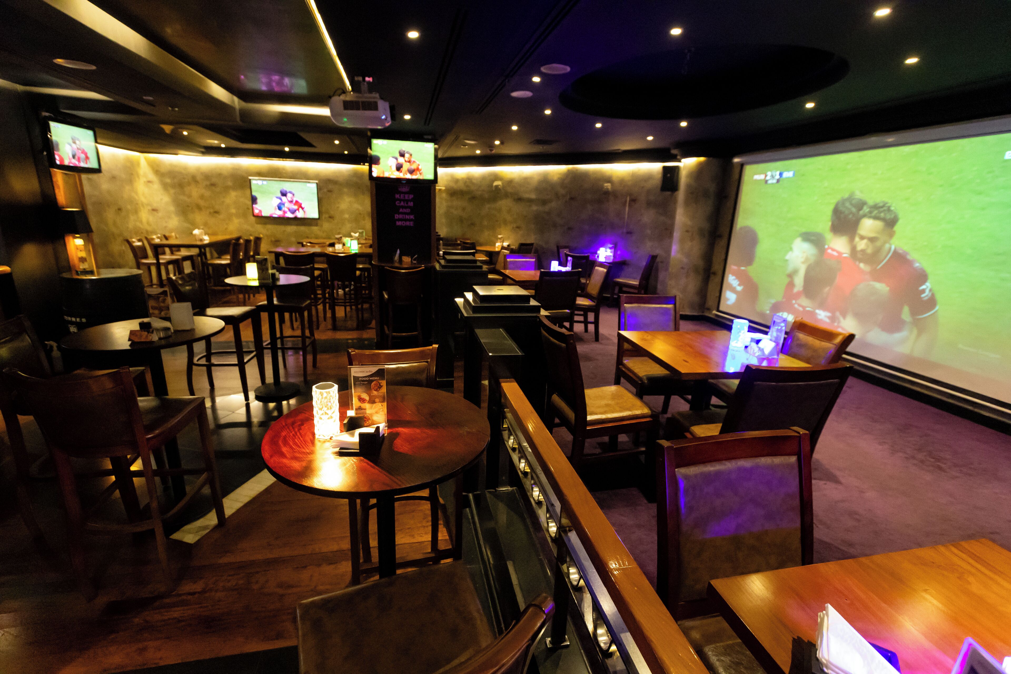 A restaurant with multiple tables and chairs, a large screen displaying a sports event, and a projector screen in the background.