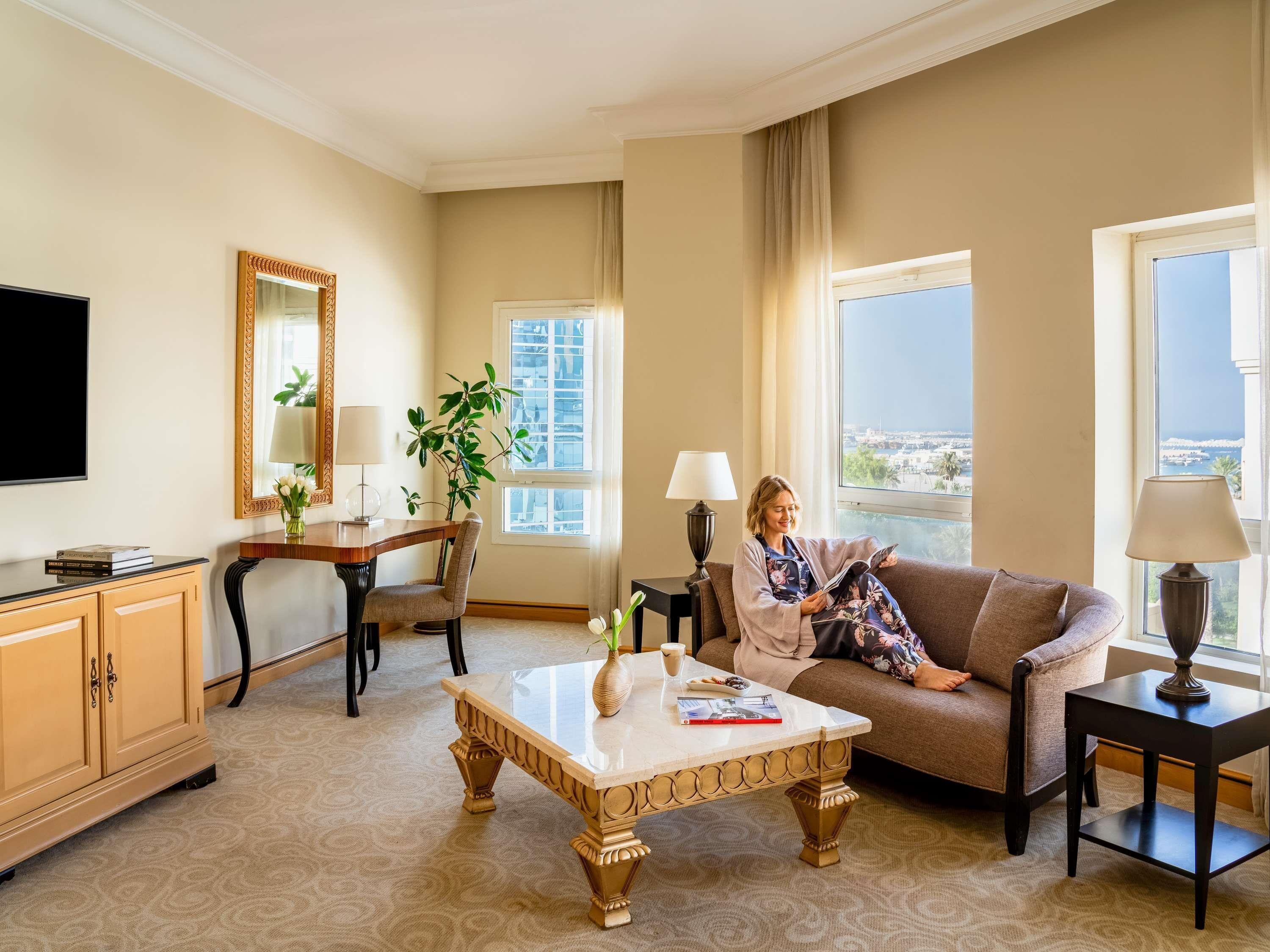 A hotel room with a large window offering a city view, a sofa, a coffee table, a TV, a desk, and a lamp.