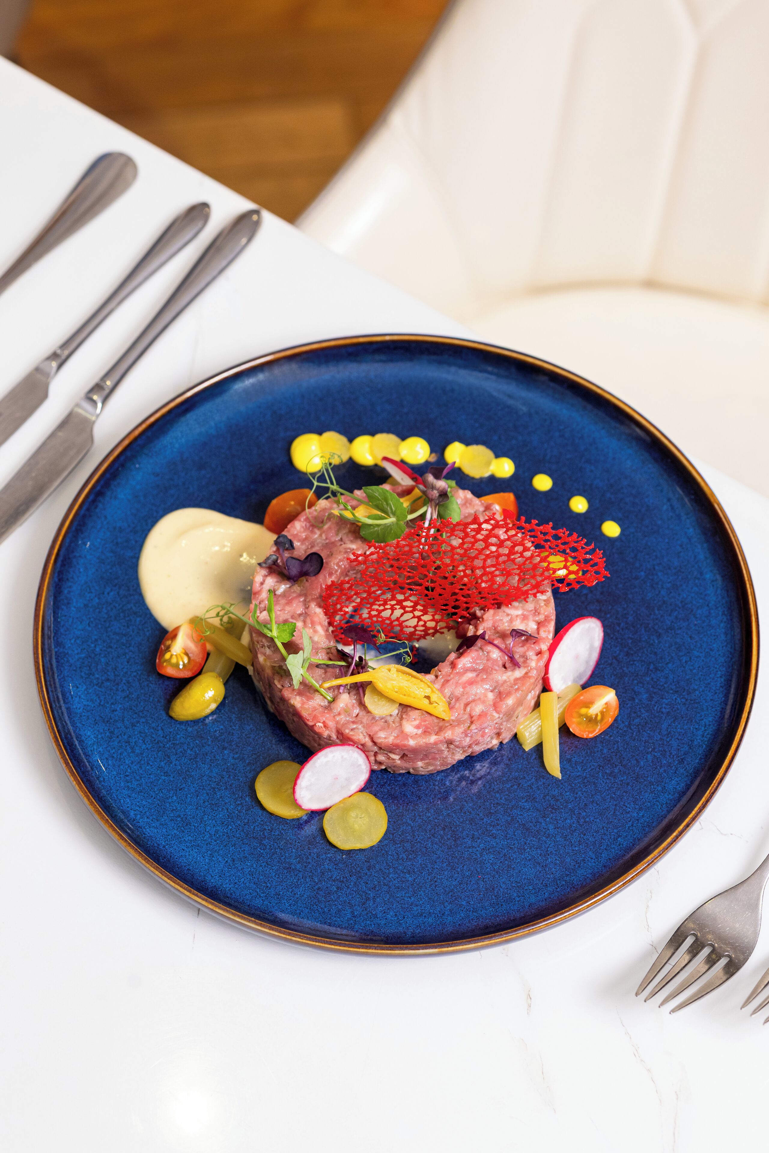 A blue plate with a steak tartare centerpiece, garnished with radishes, microgreens, and a red garnish, accompanied by a dollop of sauce and yellow dots.