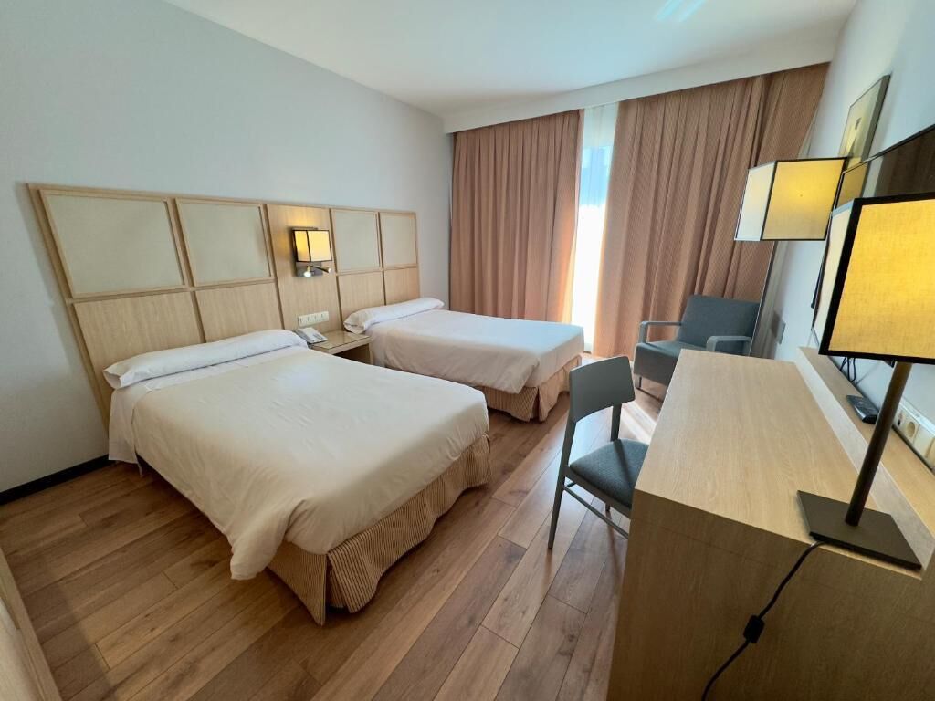 Habitación de hotel con dos camas, un escritorio, una silla y un ventanal con cortinas.