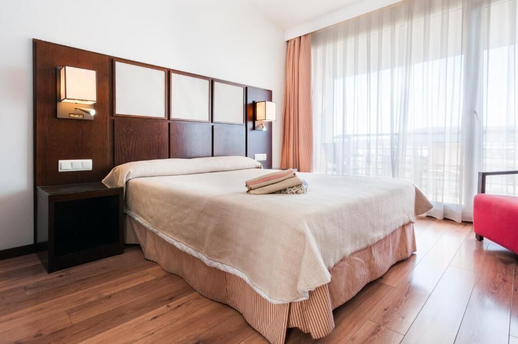 Habitación de hotel con una cama grande, cabecero de madera, lámparas de noche, una silla roja y una ventana con cortinas traslúcidas.