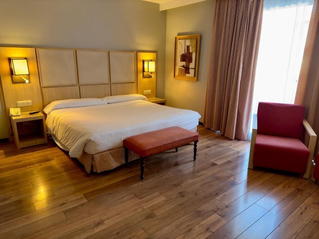 A hotel room with a large bed, bedside tables, a bench, a red chair, and a painting on the wall.