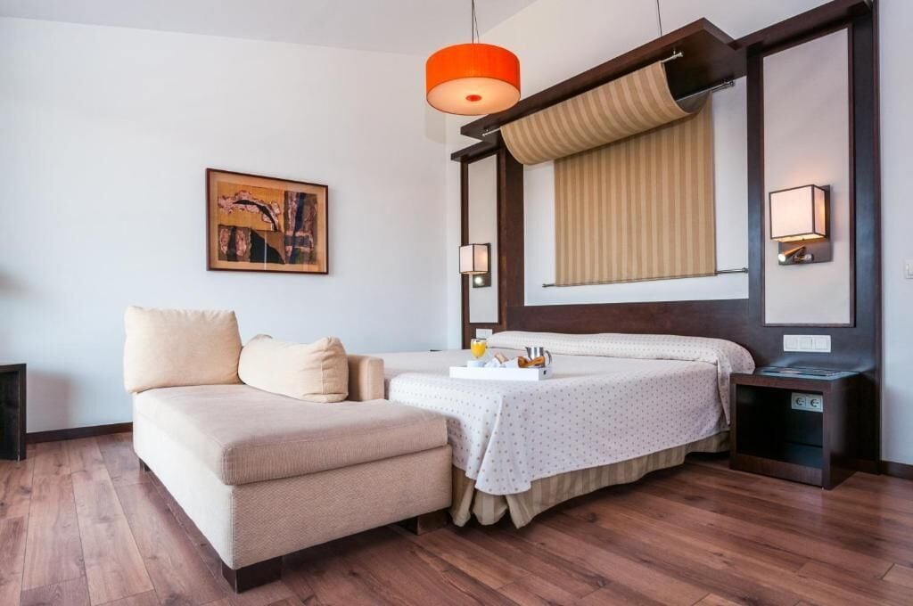 A hotel room with a bed, a sofa, a bedside table, a lamp, a painting on the wall, and a wooden floor.