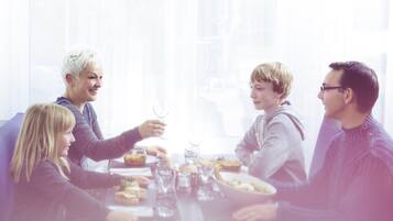 Un grupo de personas sentadas alrededor de una mesa con comida y bebidas, en una habitación luminosa con ventanas amplias.