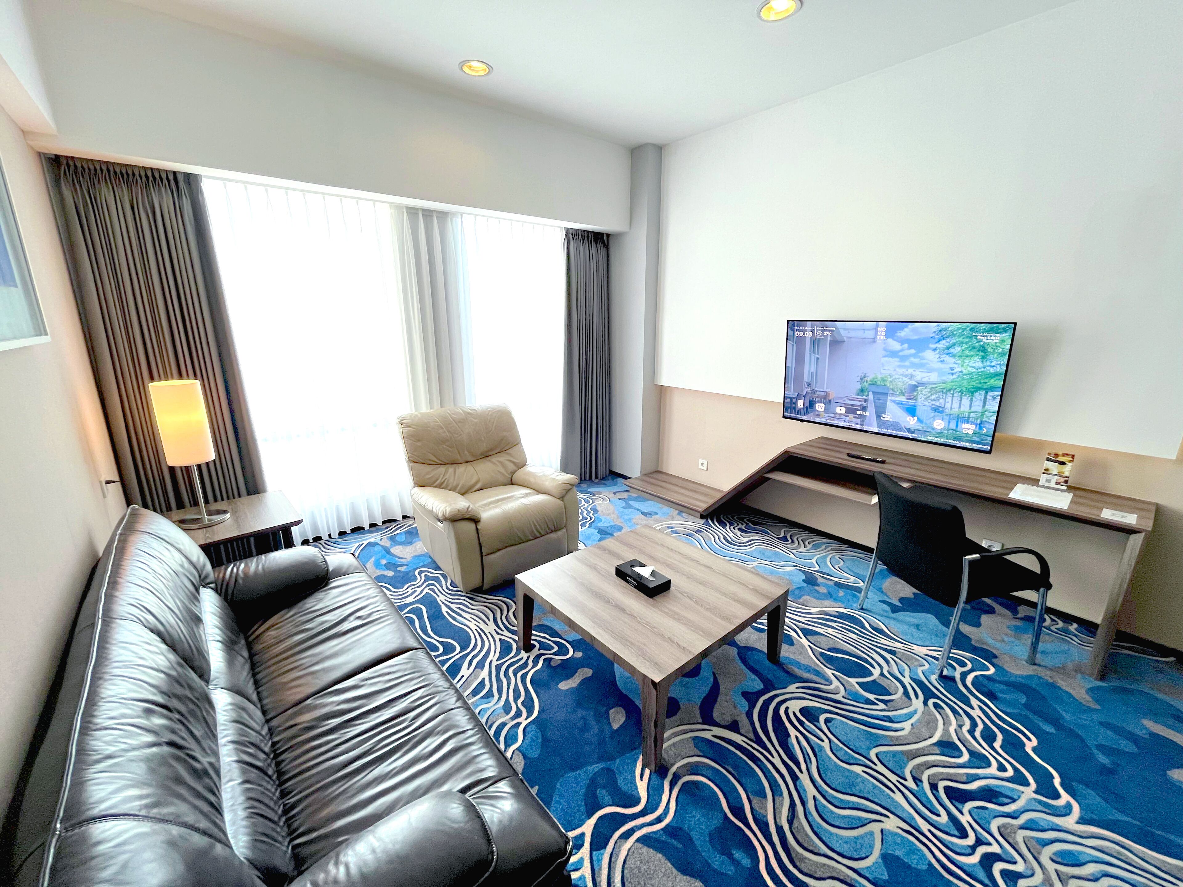 A hotel room with a sofa, a chair, a desk with a computer, a TV, and a patterned carpet.