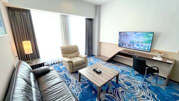 A hotel room with a sofa, a chair, a desk with a computer, a TV, and a patterned carpet.