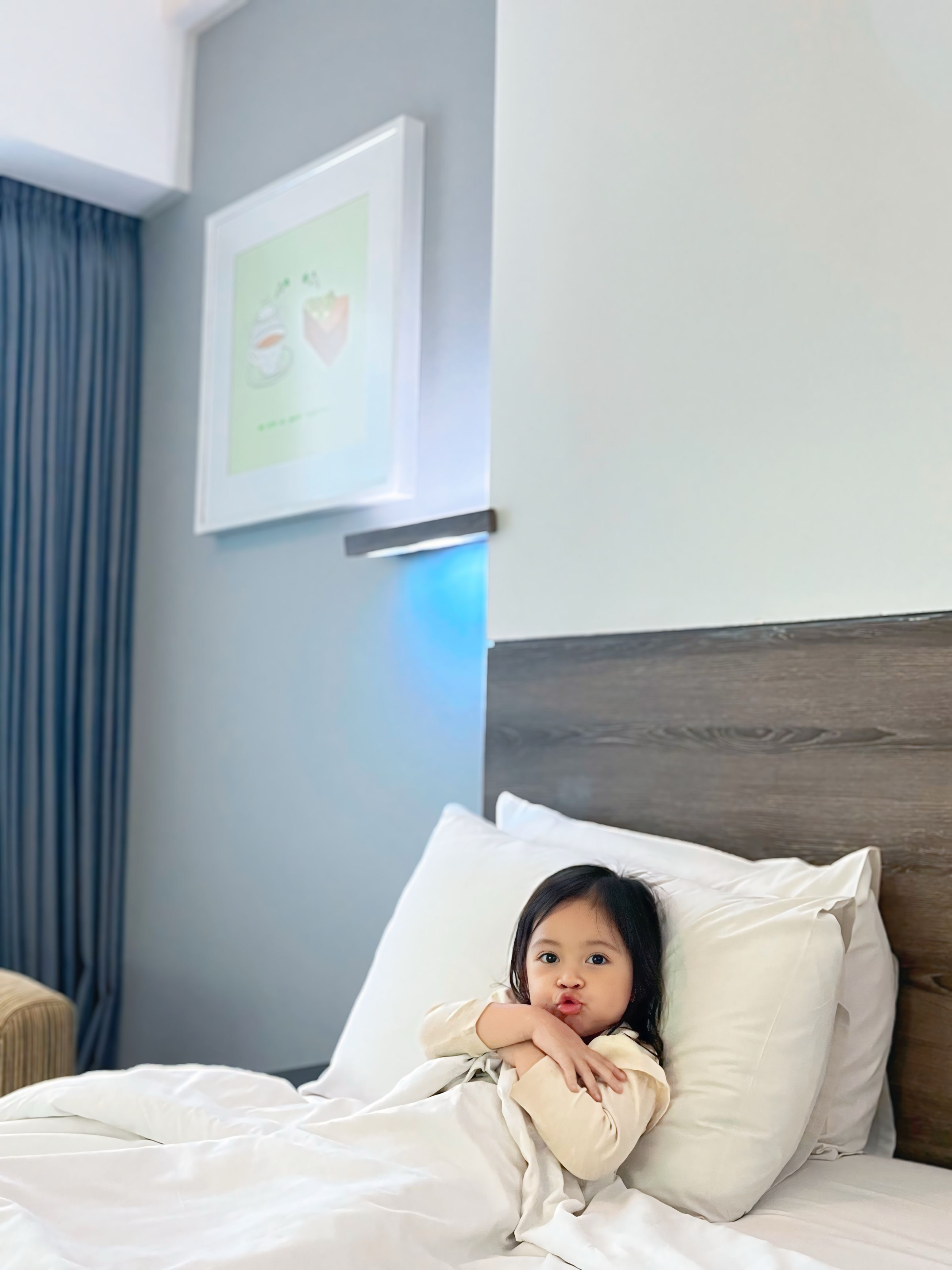 A child lying on a bed with white bedding, a wooden headboard, and a framed picture on the wall.