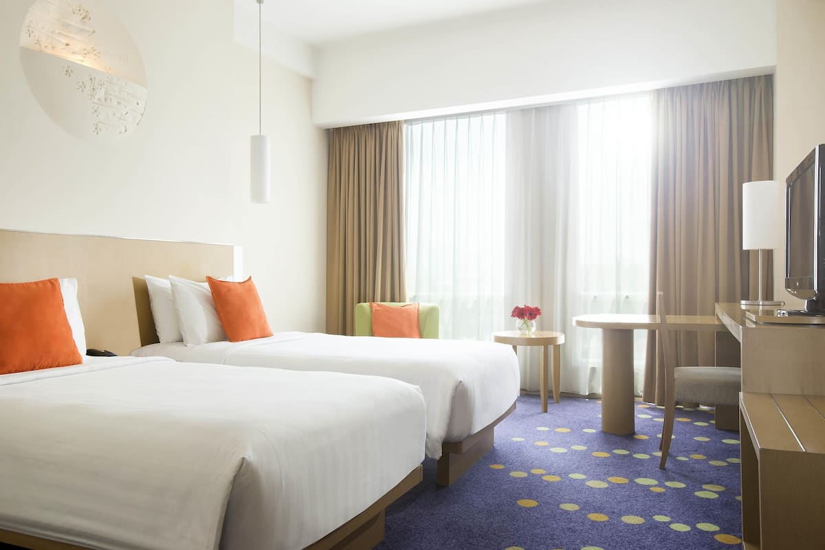 Ein Hotelzimmer mit zwei Betten, einem Schreibtisch, einem Stuhl, einem Fernseher und einem kleinen Tisch mit Blumen.