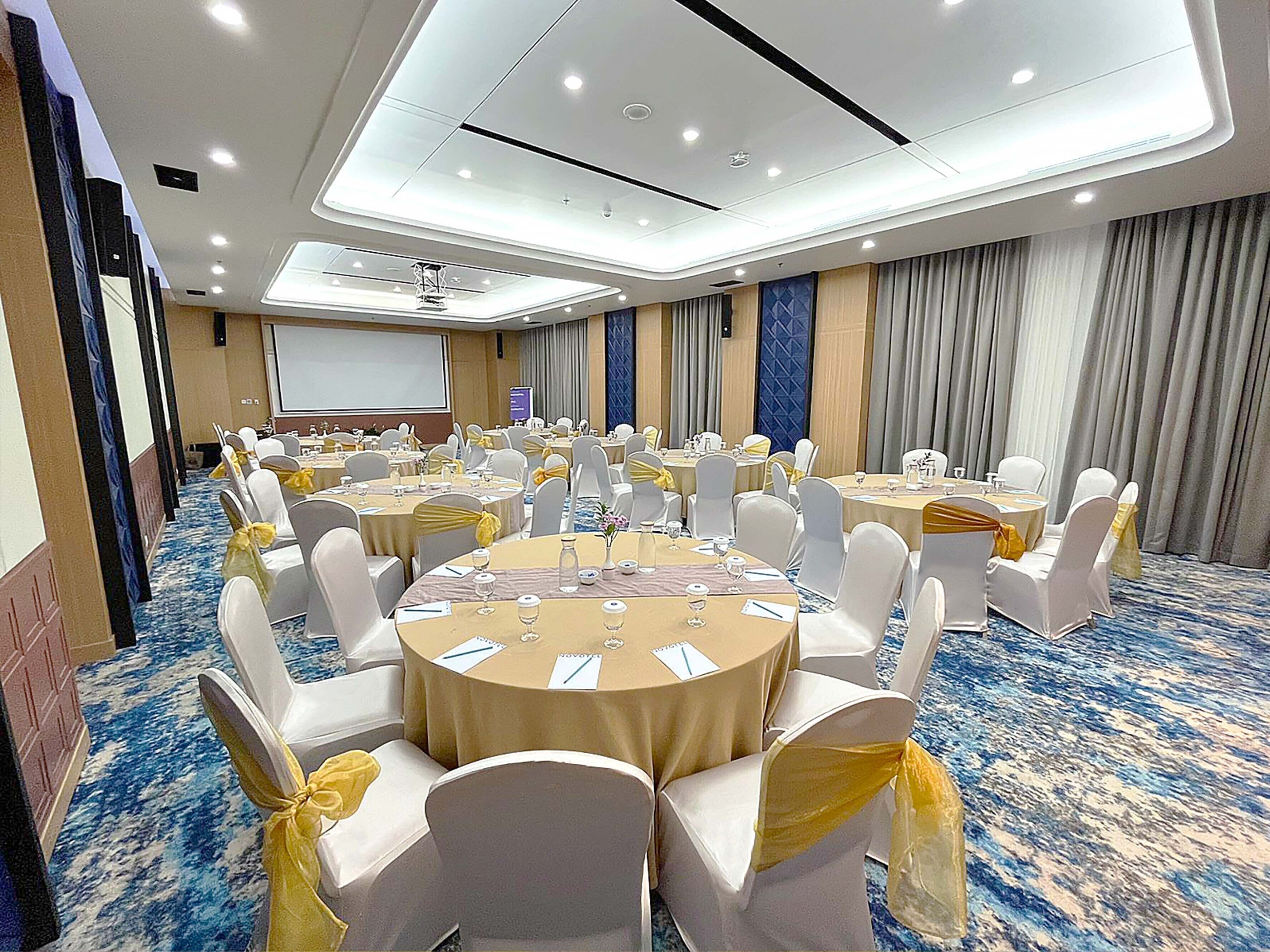 A conference room with round tables, white chairs with yellow sashes, and a projector screen.