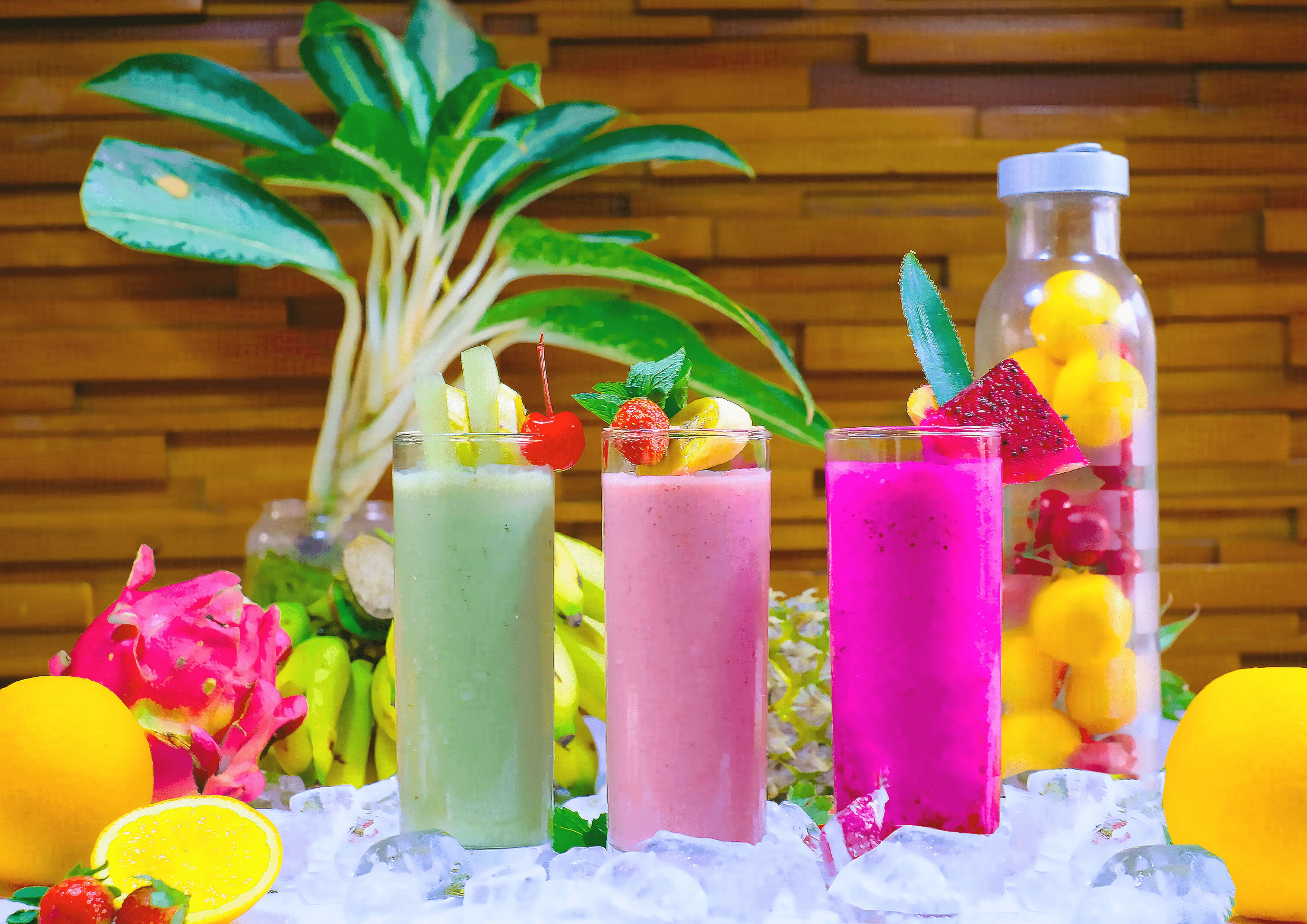 Three colorful smoothies on a bed of ice with tropical fruits and a wooden wall in the background.