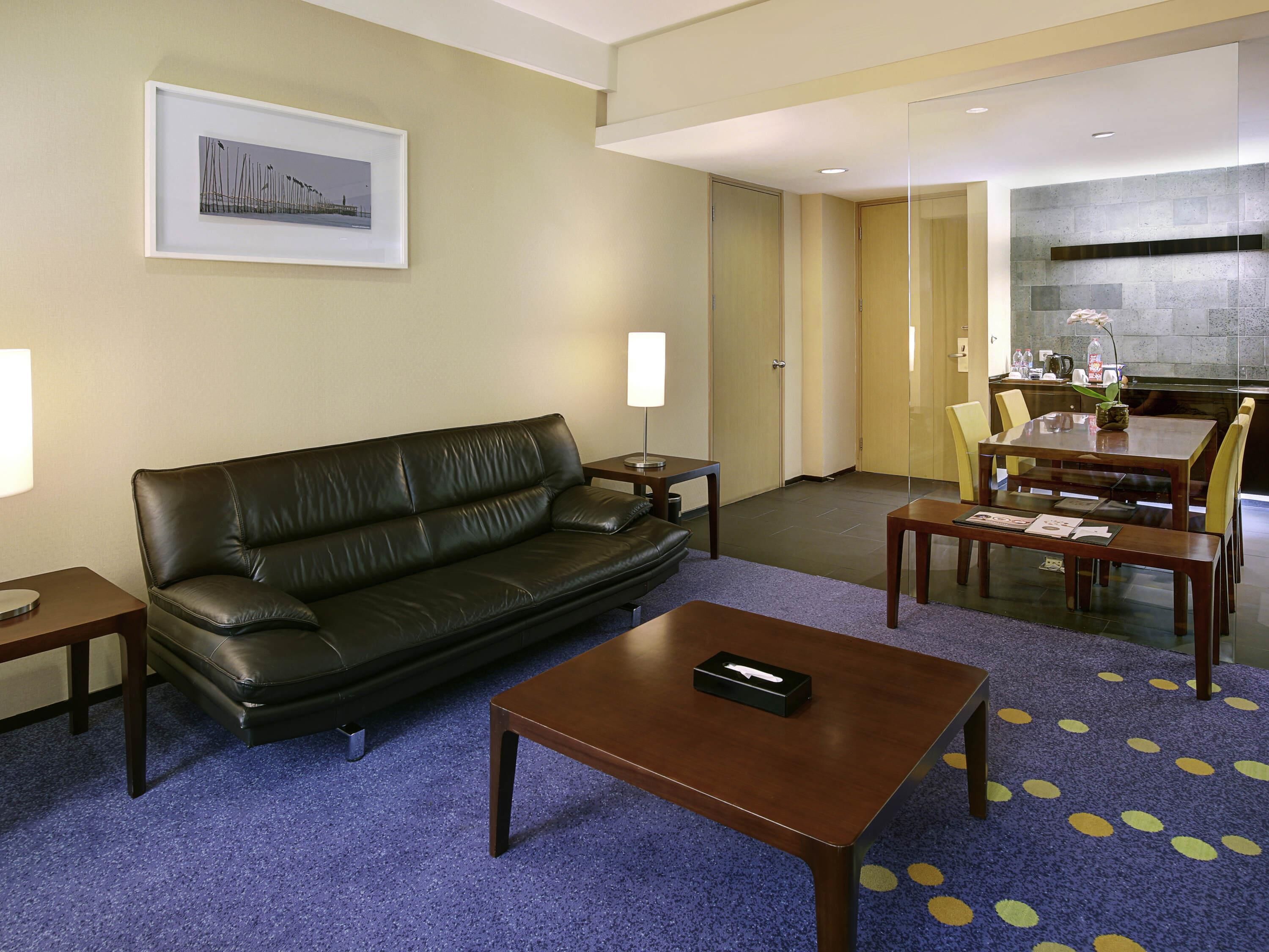 A hotel room with a leather sofa, a coffee table, a dining table with chairs, a framed picture on the wall, and a kitchen area.