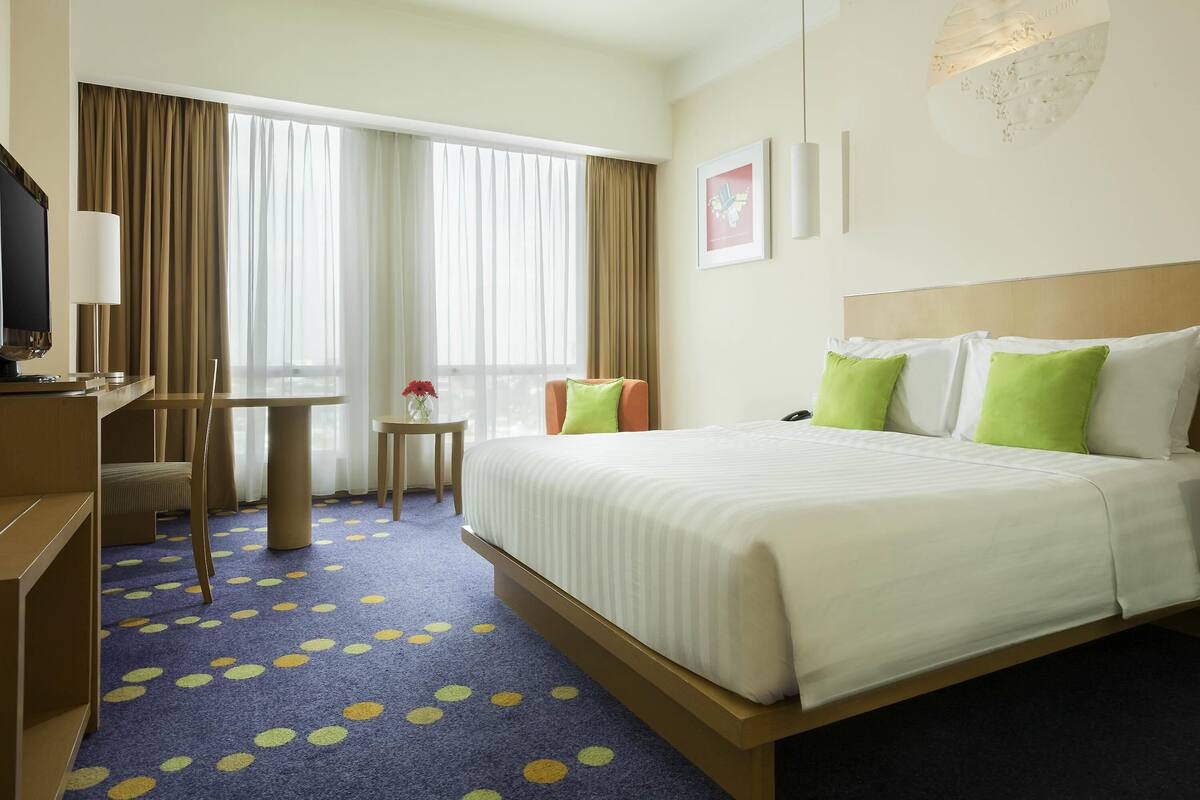 Ein Hotelzimmer mit Bett, Schreibtisch, Stuhl, einem kleinen Tisch mit Vase und großen Fenstern mit Vorhängen.