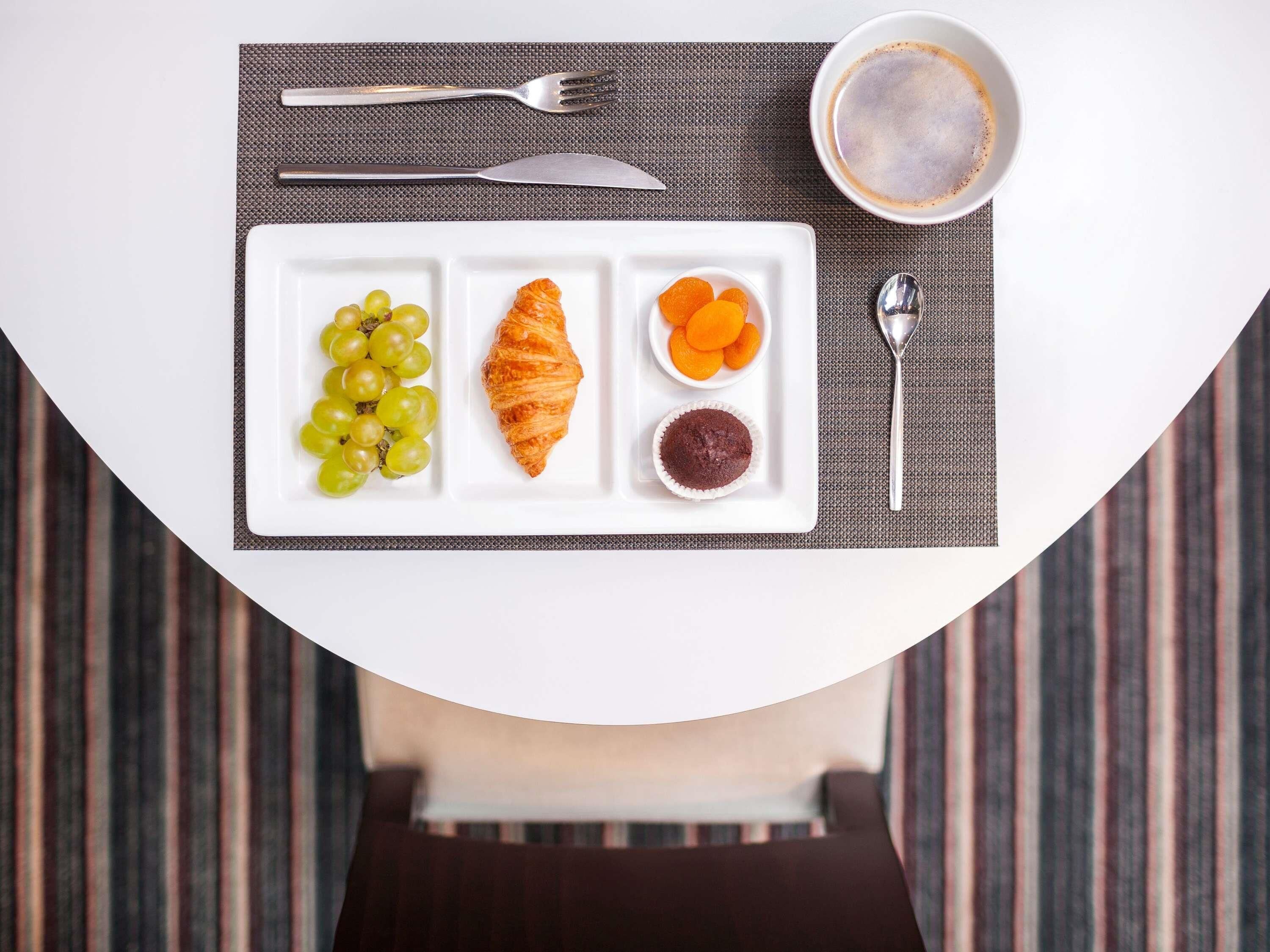 Une table dressée pour le petit-déjeuner, avec une assiette de nourriture, un croissant, des raisins et un muffin, ainsi qu’une tasse de café et des couverts.
