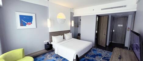 A hotel room with a bed, a desk, a chair, a bedside table, a lamp, a picture on the wall, and a door.